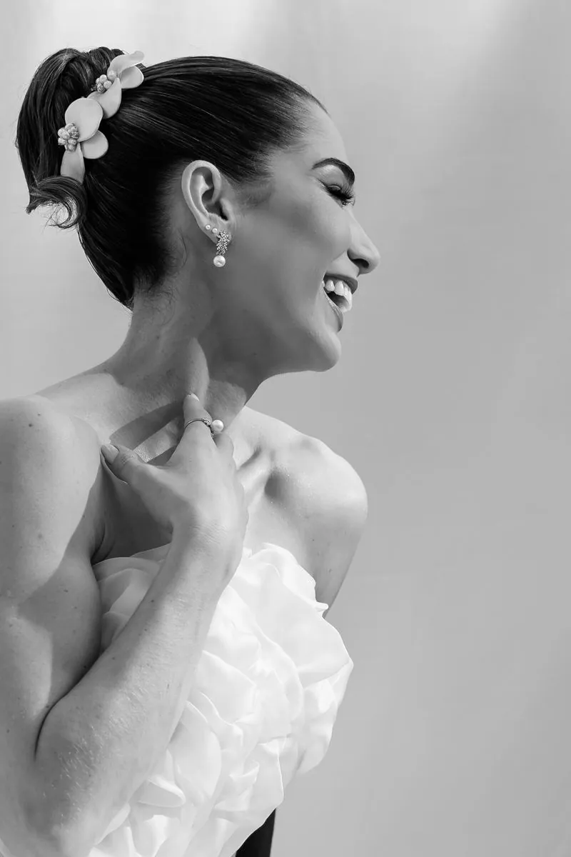 Noiva rindo de perfil, com arranjo de flores no cabelo, em preto e branco